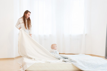 tired young mother cleaning on the house, to make a bed together with the small baby. White clothes, light house interior, family relations