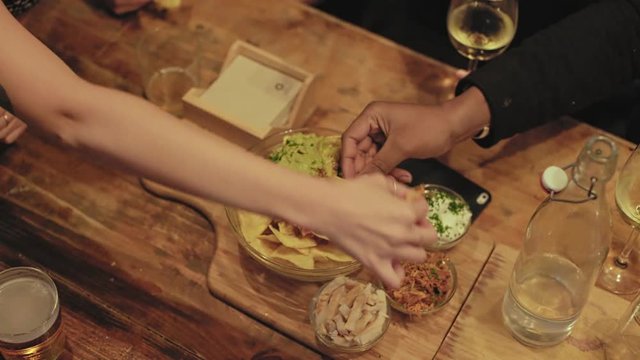 Top view of table at restaurant or bar, where group of friends share bowl of mexican nachos with guacamole and sour cream, while they drink mixed cocktails and alcohol drinks. party snacks