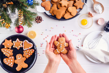 Woman hands holding traditional christmas gingerbread man cookies. White wooden table with ingrediens and decoration details. Christmas concept. The process of decorating with meringue cream.