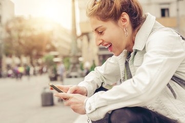 Happy woman sitting on street, typing message, chatting on smartphone
