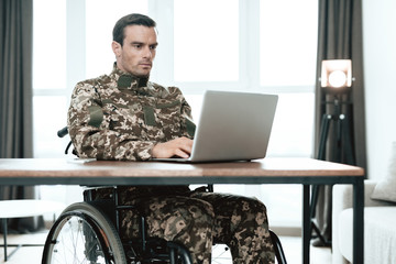 A disabled military man is sitting at a table and working with a laptop. Behind it is a large...