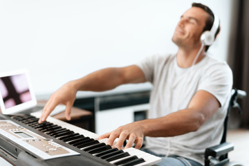 The disabled person is sitting in a wheelchair in front of the synthesizer. He plays music. He is in his large bright living room. He enjoys music.