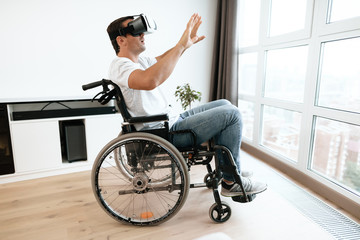 Obraz premium The disabled person is sitting in a wheelchair in virtual reality glasses. He spread his hands to the sides. He sits in his large bright living room and enjoys virtual reality.