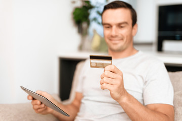 A man is sitting on the couch. He has a tablet and a credit card in his hands. He wants to buy something online. The man is smiling. He sits on a large bright sofa in his apartment.