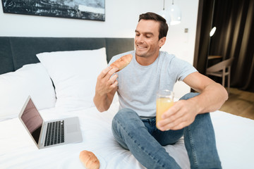 A man is sitting on the bed and having breakfast. In front of him stands an open gray laptop. He sits on a large white bed in the bedroom.