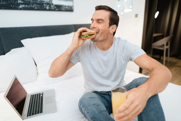 A man is sitting on the bed and having breakfast. In front of him stands an open gray laptop. He sits on a large white bed in the bedroom.