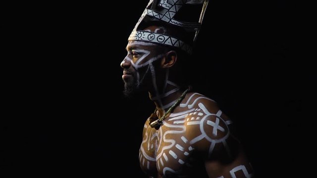 Muscular black man in white patterns smiles and dances, side view on a black background, slow motion