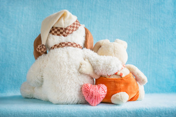 Fototapeta na wymiar Two soft toy dog and rabbit embrace and a pink knitted heart on a blue background. Two cute embracing toys puppy and bunny sitting together - girl and boy. Love, romance, tenderness, concept