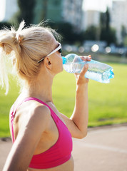 Blond slim sportswoman is drinking water from the bottle on the stadium
