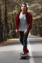 A young hipster in a cap and plaid shirt is riding his longboard on a country road in the forest