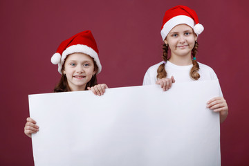 Two smiling little girl in hat of Santa Claus with white blank banner isolated on red.