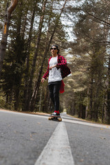 A young hipster in a cap and plaid shirt is riding his longboard on a country road in the forest