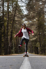 A young laughing hipster in a cap and plaid shirt is riding his longboard on a country road in the forest