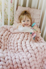 Fototapeta premium Beautiful Little Happy Blond Ginger Girl Sitting on Bed with a Pink Knitted Giant Plaid 
