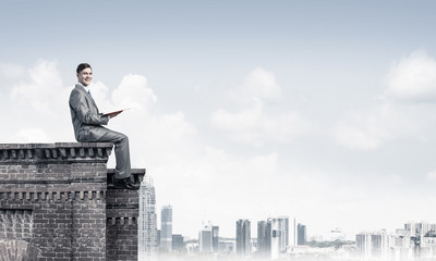 Man on roof edge reading book and cityscape at background