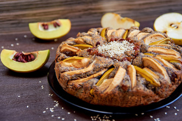 Apple pie with quince, poppy seeds, raisins and sesame on the dark plate decorated with sliced fresh apples and quince on wooden brown table. Side view 