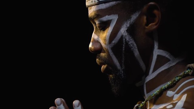 Side view as a black man in white patterns on the body with a wooden necklace around his neck praying in the dark