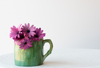 Purple daisies in green cup on white tablecloth against white background