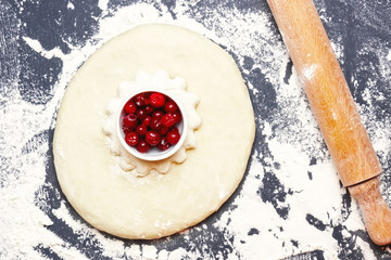 An overhead photo of sprinkled flour, wheat pie with cranberries and a rolling pin on the black table with a place for text. The process of cooking pie dough or pastry for christmas or other holidays.