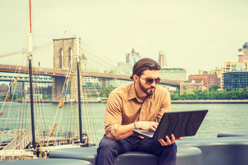 American man with beard traveling, working in New York, wearing brown shirt, sunglasses, sitting on bench by river, hunching back, reading on laptop computer. Brooklyn bridge, boat on background..