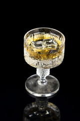 Crystal glass with yellow alcohol drink and ice cubes on a black background with reflections