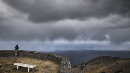 Fototapeta na wymiar Mujer asomada al mirador de Vixía Herbeira ve acercarse la tormenta, Costa ártabra, cerca de san Andrés, Galicia, españa