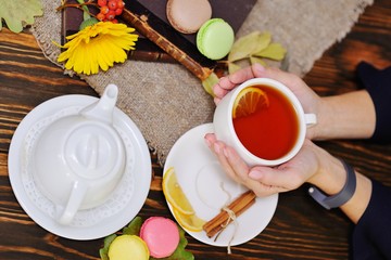 women's hands hold a cup of tea with lemon on a wooden background and against a backdrop of macaroons, old books and burlap
