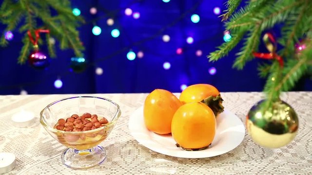 Persimmon, Nuts with honey and are on the table with a decorated Christmas tree and flashing gerunds blurry bokeh. Photographed with dolly from right to left