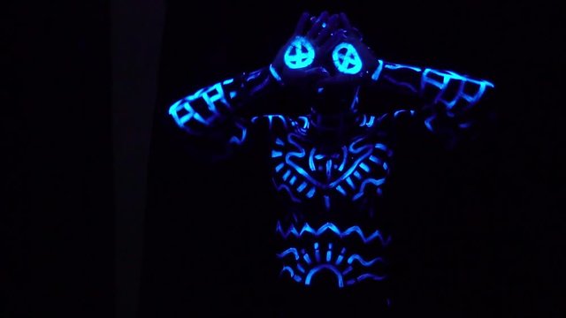 Conceptual shooting of a black man in neon patterns on his body that stands in the dark and shows a thumbs-up and covers his face with his hands, slow motion