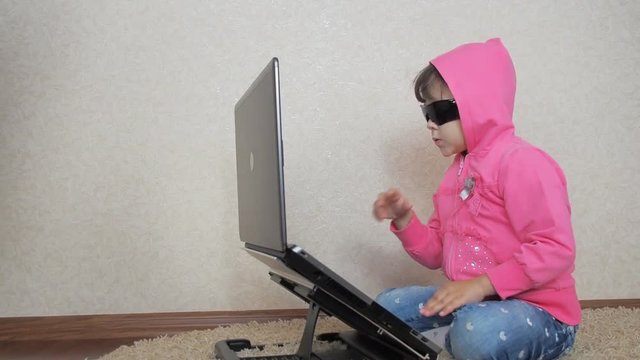 A little hacker. A child wearing sunglasses and a hood with a laptop.