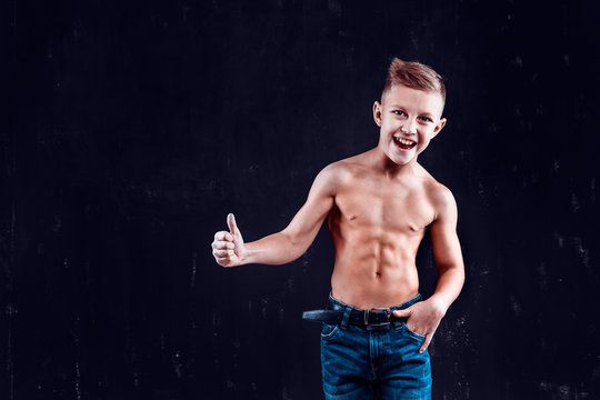 Happy bare-chested boy in jeans showing thumb-up gesture while standing on dark wall background.