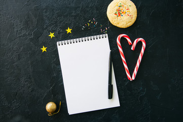 Festive background, white empty page of notepad, candy canes, cookie with confetti, stars on a black stone background. Top view with copy space. Flat lay.
