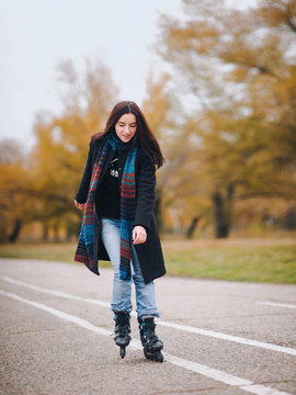A pretty girl in a coat and with a long scarf happily skates in the autumn park. Fighting the autumn depression. To overcome depression.