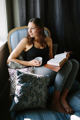 Obraz premium Happy woman sitting in a chair and looking out the window with her coffee and book