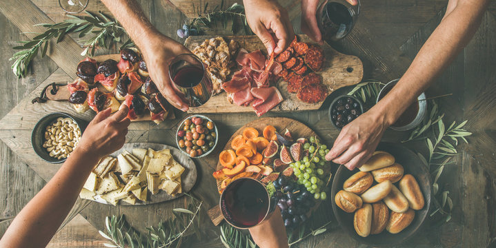 Flat-lay of friends eating and drinking together. Top view of people having party, gathering, celebrating at wooden rustic table set with various wine snacks and fingerfoods. Hands holding glasses