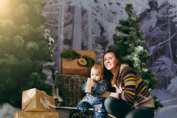 Young playful happy mother with a cute little baby boy in a decorated forest with fir trees, sledges and gift boxes. Christmas good mood. New Year. Lifestyle, family and togetherness concept.
