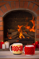 Hot drinks in mugs on wooden table beside cosy open fire place. Autumn or winter holidays concept,...