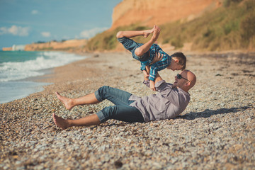 Touching appealing scene of father and son enjoy summer vacation together playing on stone beach wearing stylish shirt and fashion blue jeans both barefoot with adore landscape on background
