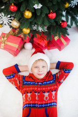 Portrait of cute funny little kid dressed in red pajamas and hat of santa. White happy kid laying on white wooden floor near holiday presents and tree. He smiles happily. Flat lay vertical color image