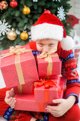 Fototapeta na wymiar Portrait of cute funny little kid dressed in red night-suit and hat of santa. White happy kid holding stack of holiday presents in hand and smiles happily. Vertical color photo.