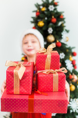 Obraz na płótnie Canvas Portrait of cute funny little kid dressed in red night pajamas and hat of santa. Blurry white happy kid holding pile of holiday presents in hands and smiles happily. Focus at foregraund.