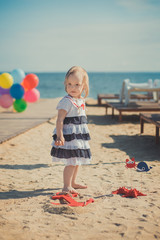 Cute blond baby girl child posing enjoying summer life time on sandy beach sea side on wooden pier with colourful plenty balloons wearing casual dress barefoot