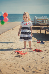 Cute blond baby girl child posing enjoying summer life time on sandy beach sea side on wooden pier with colourful plenty balloons wearing casual dress barefoot