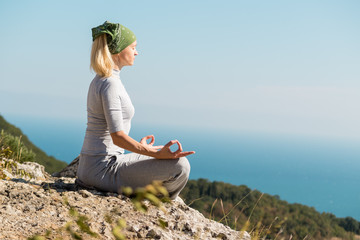 Beautiful blond yoga woman sitting on the top of the mountain in lotus pose. Meditation on the edge with a scenic view of the landscape and the sea.