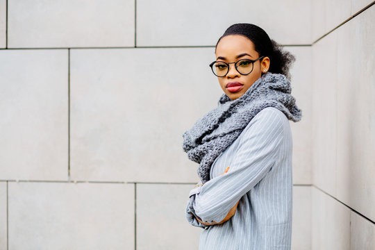 Side view of portrait beautiful african american business woman in glasses and gray kniitted snud smiling and looking at camera over beige wall. Lifestyle, leisure and people concept.