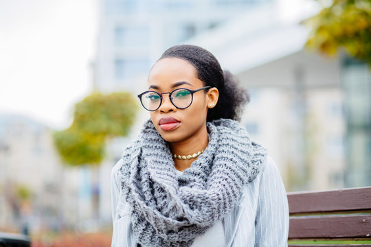 Portrat Of Serious Nigerian Student Business Woman In Glasses, Knitted Scarf And Suit Sitting On The Bench Over Urban City Background. Lifestyle, Leisure And People Concept.