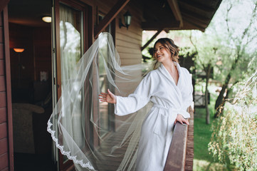 The bride stands in a white robe in the morning before the wedding, throwing a veil on her shoulders.