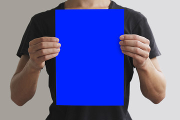Man in black t shirt holding blank blue A4 paper vertically. Leaflet presentation. Pamphlet hold hands. Man show clear offset paper. Sheet template. Isolated on grey background