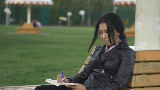 Beautiful young sad woman in black leather jacket sitting on bench in the park and writing something with pen in her personal diary