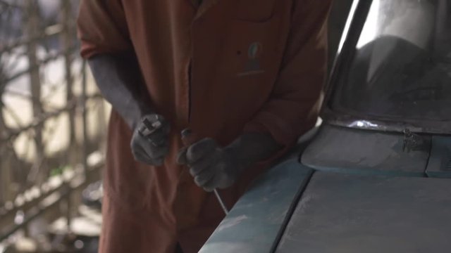 African mechanic chips off paint  from vehicle with hammer and chisel.